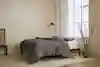 A minimalist bedroom with a bed featuring gray bedding, a wooden frame, a black floor lamp, a large window, a ceramic vase with branches, and neutral decor on a light carpeted floor.