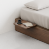 A minimalist wooden bed with a built-in side tray holding a black coffee cup and saucer, a closed notebook, and a pen, set against a light-colored wall and floor.