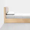 A minimalist wooden bed with a light wood frame, a cushioned headboard, white bedding, a pillow, and neatly arranged sheets against a plain white wall and concrete floor.