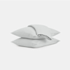 Two neatly stacked light gray pillows, with a folded matching bedsheet or duvet placed on top, set against a plain white background.