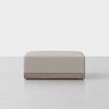 A modern, rectangular ottoman with light beige upholstery and a wooden base, placed on a smooth, light-colored floor against a plain white background.