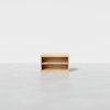 A small, light wood rectangular shelf unit with one open shelf, placed on a plain light gray floor against a white background.