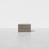 A minimalist wooden nightstand with two drawers, placed in the center of a light gray, empty room with a plain white background.