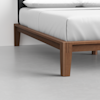 A close-up view of a modern wooden bed frame with clean lines, supporting a mattress with white bedding, set on a light gray floor against a plain white wall.