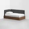 A modern daybed with a wooden frame and dark gray upholstered backrest is placed in the corner of a minimal, white-walled room. The bed is neatly made with white bedding and a single pillow.
