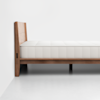 A side view of a minimalist wooden bed frame with a white mattress on top, set against a plain white wall and floor.