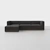 A modern, dark gray L-shaped sectional sofa with low backrests and a wooden base, placed on a light gray floor against a plain white background.