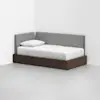 A modern daybed with a dark wooden frame and gray upholstered backrest sits in the corner of a white room. The bed is made with white bedding and a single pillow.