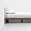 A minimalist bed with a light gray upholstered headboard, white bedding, and a wooden frame, set against a plain white wall and floor.