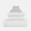 A neatly stacked set of bedding, including two pillows, a comforter, and several folded sheets and pillowcases, all in white and light gray, arranged in a pyramid shape against a plain white background.