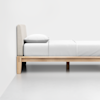 A minimalist wooden bed frame with a beige upholstered headboard, topped with white bedding and a pillow, stands against a plain white wall on a smooth, light gray floor.