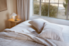 A sunlit bedroom with a neatly made bed, two pillows, an open book, and a glass of water on a wooden nightstand beside a window with beige curtains overlooking a garden.