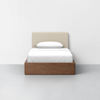 A single bed with a beige upholstered headboard, white bedding, and a wooden frame, placed against a plain white wall on a light gray floor.