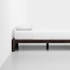 A minimalist bedroom with a single wooden bed frame, a neatly made white mattress, and a white pillow, placed against a plain white wall on a smooth, light-colored floor.