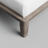 Close-up view of a wooden bed frame corner with a white mattress on top, showcasing the smooth finish and detailed joinery of the leg against a light gray floor.
