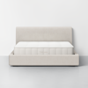 A minimalist beige upholstered bed frame with a matching headboard and a white mattress, set against a plain white wall and gray floor. The word THUMA is visible on the side of the mattress.