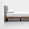A minimalist bed with a wooden frame, gray upholstered headboard, and white bedding, including a single pillow, placed against a plain white wall on a light floor.
