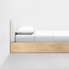 A minimalist wooden bed frame with a padded beige headboard, white bedding, and a single pillow, placed against a plain white wall on a gray floor.