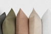 Five textured throw pillows in gray, olive green, dusty pink, beige, and white are arranged upright in a row against a plain white background.