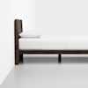 A modern, dark wood bed frame with a white mattress, white bedding, and a single white pillow, positioned against a white wall and set on a light-colored floor.