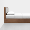 Side view of a minimalist wooden bed with a padded headboard, white pillow, and white bedding, set against a plain white wall and gray floor.
