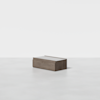 A small, rectangular wooden storage box with a flat lid sits on a plain, light-colored floor against a white background.