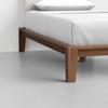 A close-up view of the corner of a wooden bed frame with clean lines, supporting a mattress with white bedding on a light gray floor.