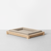 A minimalist pet bed with a light wood frame and a soft, rectangular beige cushion, placed on a smooth gray floor against a plain white background.