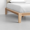 Close-up of a wooden bed frame corner with a white mattress and neatly tucked white bedding, set on a light gray floor against a plain white wall.