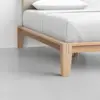 Close-up of a wooden bed frame corner with a white mattress and neatly tucked white bedding, set on a light gray floor against a plain white wall.
