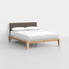 A modern bed with a light wood frame, white bedding, two white pillows, and a dark upholstered headboard, set against a plain white wall and floor.