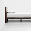 Side view of a minimalist wooden bed with a light-colored upholstered headboard, white bedding, and a white pillow, positioned against a plain white wall in a bright room.