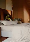 A cozy bedroom with a neatly made bed, beige sheets, and pillows. A light blanket is draped over the bed. Warm wooden walls, houseplants, and soft natural lighting create a calm, inviting atmosphere.