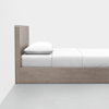 A minimalist wooden bed frame with a headboard, fitted with a white mattress, white pillow, and white bedding, placed against a plain white wall on a light gray floor.