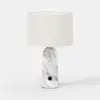 A modern table lamp with a cylindrical white fabric shade and a marble-patterned base featuring a small black circular accent near the bottom. The background is plain white.
