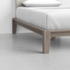 A close-up view of a bed with a light wood frame and white bedding, positioned on a smooth, light gray floor in a minimal, modern room.