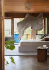 A white sheet floats in midair above an unmade bed in a modern, sunlit bedroom with wood accents, large windows, and a leafy view outside. A small table with a candle sits beside the bed.