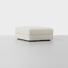 A modern, square, low-profile ottoman upholstered in light beige fabric sits on a smooth, light gray floor against a plain white background.