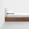 A minimalist bed with a light upholstered headboard, white pillow, and white bedding sits against a white wall. The bed frame is made of wood, and the room has a light gray floor.