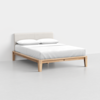 A modern bed with a light wooden frame, white mattress, white bedding, and two white pillows, set against a plain white wall and a light-colored floor.