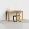 A minimalist wooden desk with a matching chair sits on a light rug. The desk holds a small lamp and an open notebook, set against a plain white wall in a bright, uncluttered room.