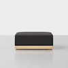 A modern, square ottoman with a black cushioned top and a light wood base, placed on a light gray surface against a plain white background.