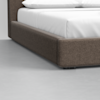 Close-up view of the corner of a modern bed with a gray upholstered frame and white bedding, set on a light gray floor.