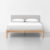 A modern bed with a light wood frame, grey upholstered headboard, and white bedding, including two pillows, is placed in a minimalist room with a plain white wall and a light gray floor.