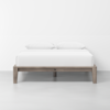A minimalist wooden bed frame with a light wood finish, neatly made with white bedding and two white pillows, set against a plain white wall in a clean, modern room.