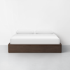 A modern bed with a dark wooden frame, white bedding, and two white pillows, set against a plain white wall and a light gray floor.