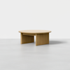 A minimalist wooden coffee table with a round top and three flat, rectangular legs, displayed in a bright, empty space with a light gray floor and plain white background.