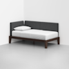 A modern daybed with a dark wooden frame, white mattress, single white pillow, and dark gray upholstered back and side cushions, placed in the corner of a minimalist white room.