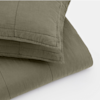A folded olive green quilted blanket with matching pillow shams, both featuring visible stitching and a soft, textured fabric, displayed against a plain white background.