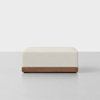A square, cream-colored ottoman with a soft fabric upholstery and a wooden base, placed on a light gray floor against a plain light background.